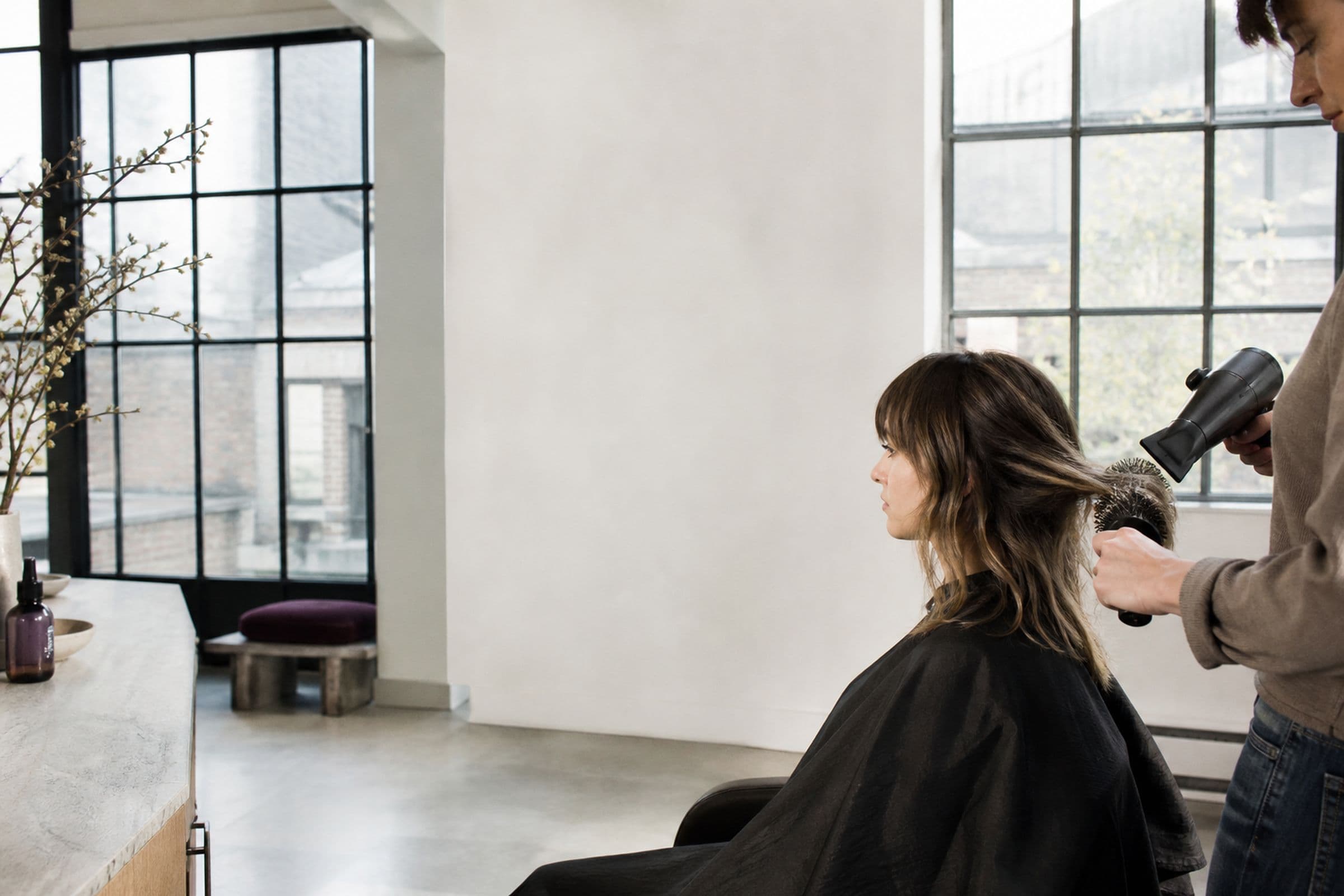 A stylist blow-drying a client’s hair in soft light from tall windows